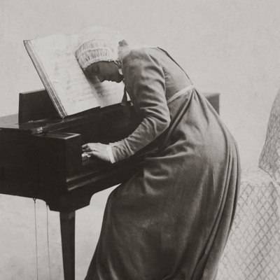 Schwarzweißaufnahme einer Frau in altertümlicher Kleidung vor einem Cembalo oder Klavier.